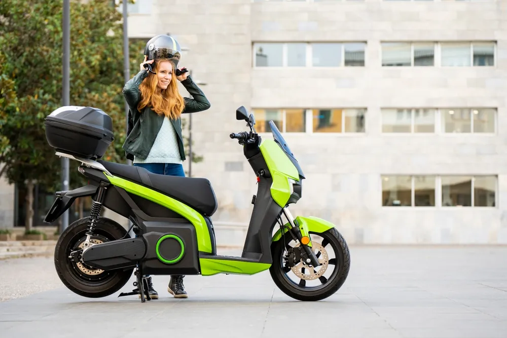 Mujer con cabello rizado y largo, usando chaqueta negra, colocando un casco sobre su cabeza junto a una moto eléctrica verde.