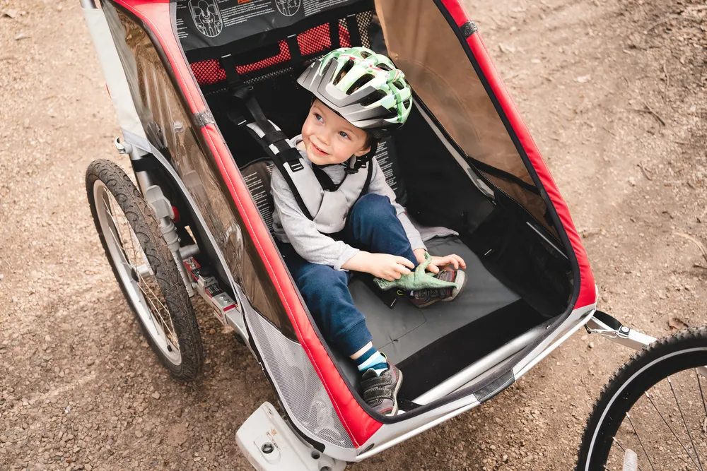 Niño pequeño sentado en un remolque de bicicleta, usando casco y jugando con un juguete, en un camino de tierra.