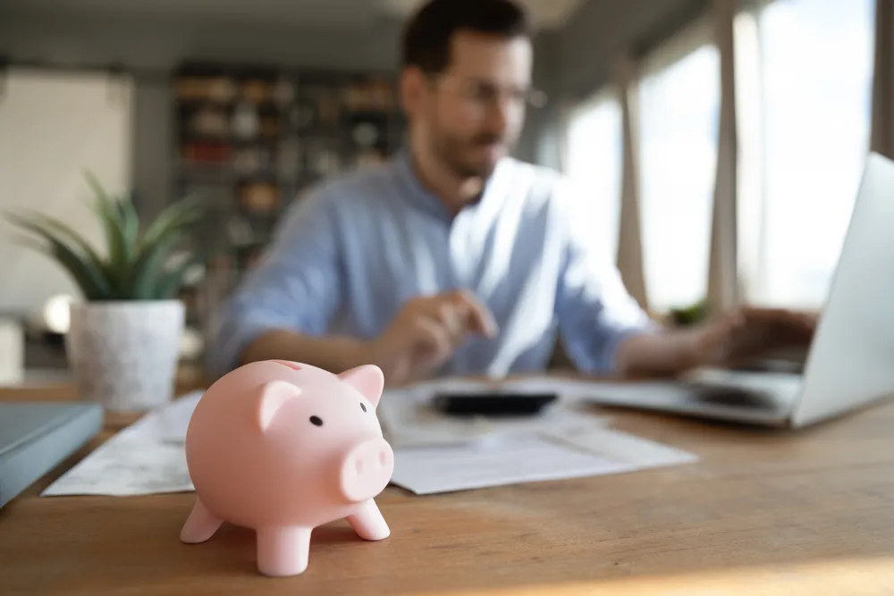 Cerdito de ahorro rosa en primer plano, con un hombre trabajando en una laptop y documentos en una mesa de madera.