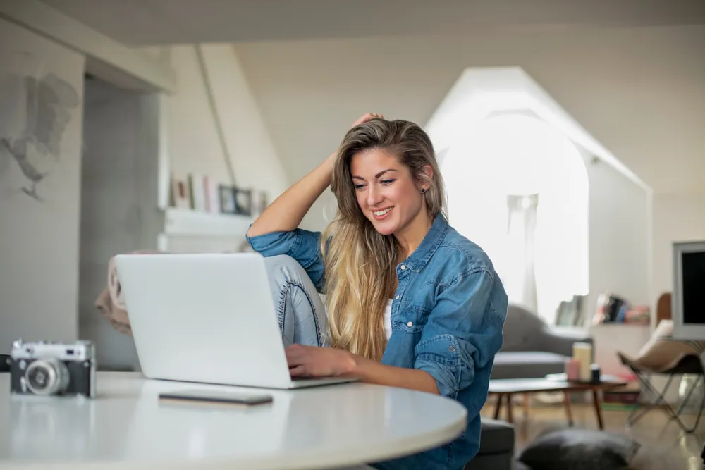 Mujer sonriente con cabello largo, usando una laptop en una mesa, en un ambiente luminoso y moderno.