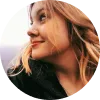 Mujer con cabello rubio y ondulado, sonriendo mientras mira hacia un lado, fondo desenfocado.