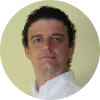 Hombre con cabello rizado y camisa blanca, posando frente a un fondo amarillo.