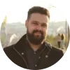 Hombre con barba y cabello corto, sonriendo, vestido con chaqueta negra, fondo natural iluminado.