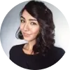 Mujer con cabello oscuro y rizado, sonriendo, vestida con una camiseta negra, fondo claro.