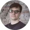Hombre con gafas y cabello oscuro, frente a un fondo de ladrillos expuestos.