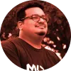 Hombre sonriente con gafas, cabello corto y barba, vestido con camiseta negra, fondo en tonos rojos.