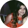 Mujer con cabello largo y ondulado, vistiendo una blusa naranja y un collar, sonríe en un entorno oscuro.