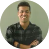 Hombre sonriente con camisa de cuadros, brazos cruzados, fondo claro y circular.