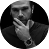 Hombre con barba y reloj, posando con una mano en la cara, fondo oscuro.