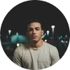 Joven con camiseta clara, mirando a la cámara, fondo oscuro con luces difusas.
