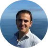 Hombre sonriente con camisa clara, fondo de mar y cielo despejado.