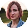 Mujer con cabello corto y rosa, mirada seria, vestida con chaqueta de mezclilla, fondo natural desenfocado.