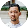 Hombre joven con cabello oscuro y barba ligera, viste una camisa blanca con puntos, sonríe frente a un fondo urbano.
