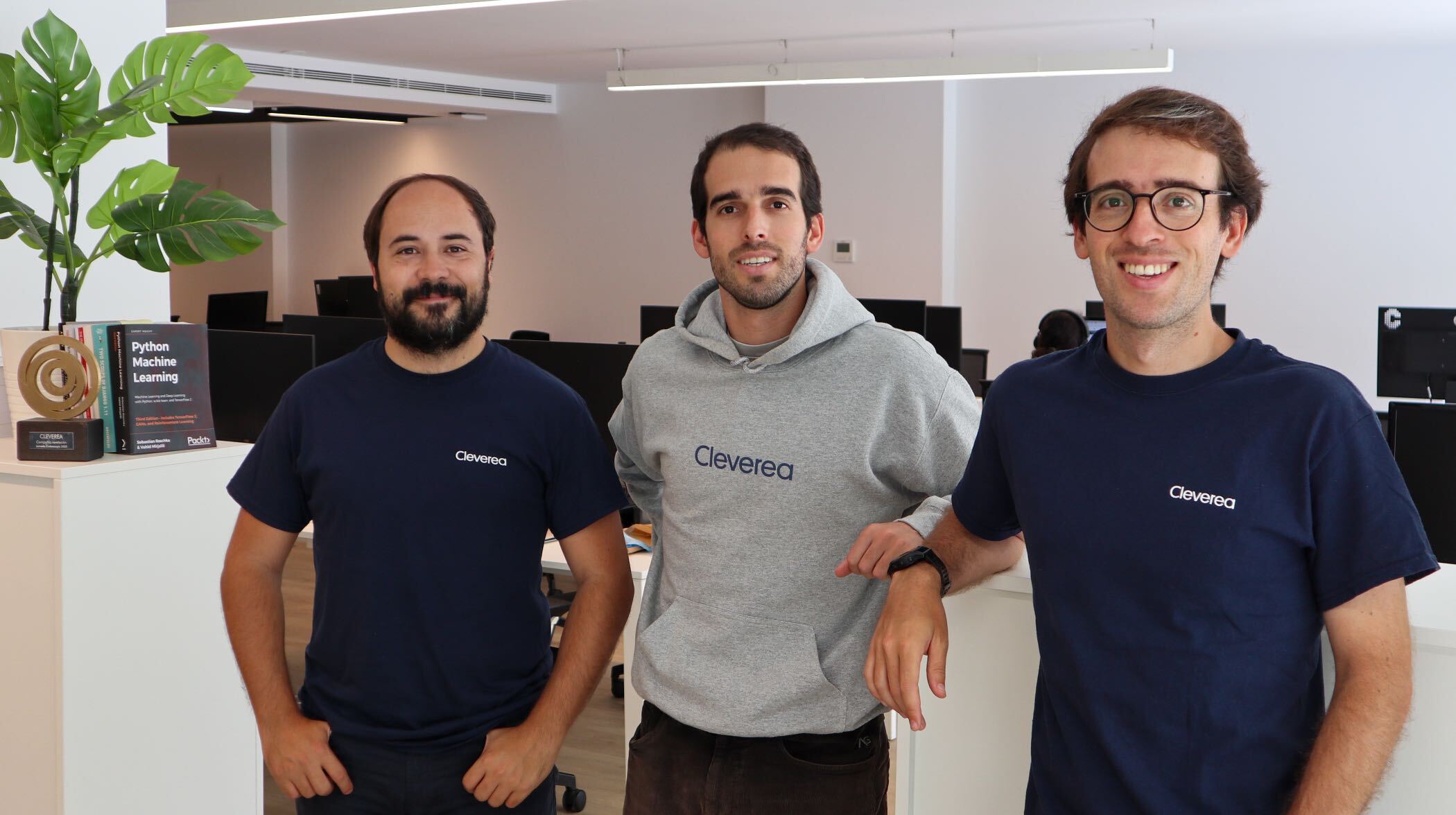 Javier, Joan y Álvaro, fundadores de Cleverea, sonrientes en un entorno de oficina moderna, con plantas y computadoras de fondo.