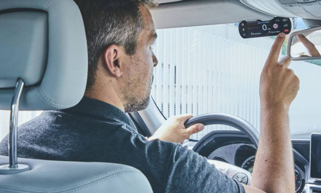 Hombre sentado al volante de un coche, ajustando un dispositivo en el espejo retrovisor.
