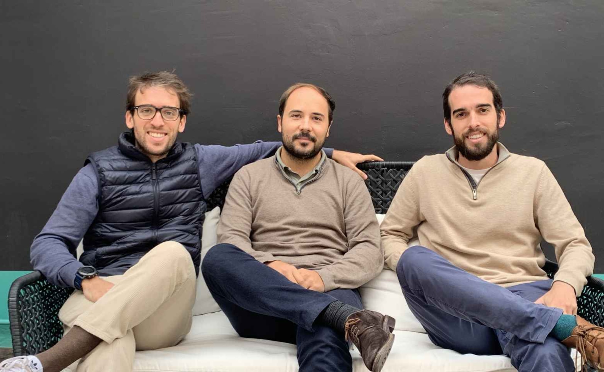 Javier, Joan y Álvaro, fundadores de Cleverea,sentados en un sofá, sonriendo. Dos llevan suéteres claros y uno un chaleco negro. Fondo oscuro.