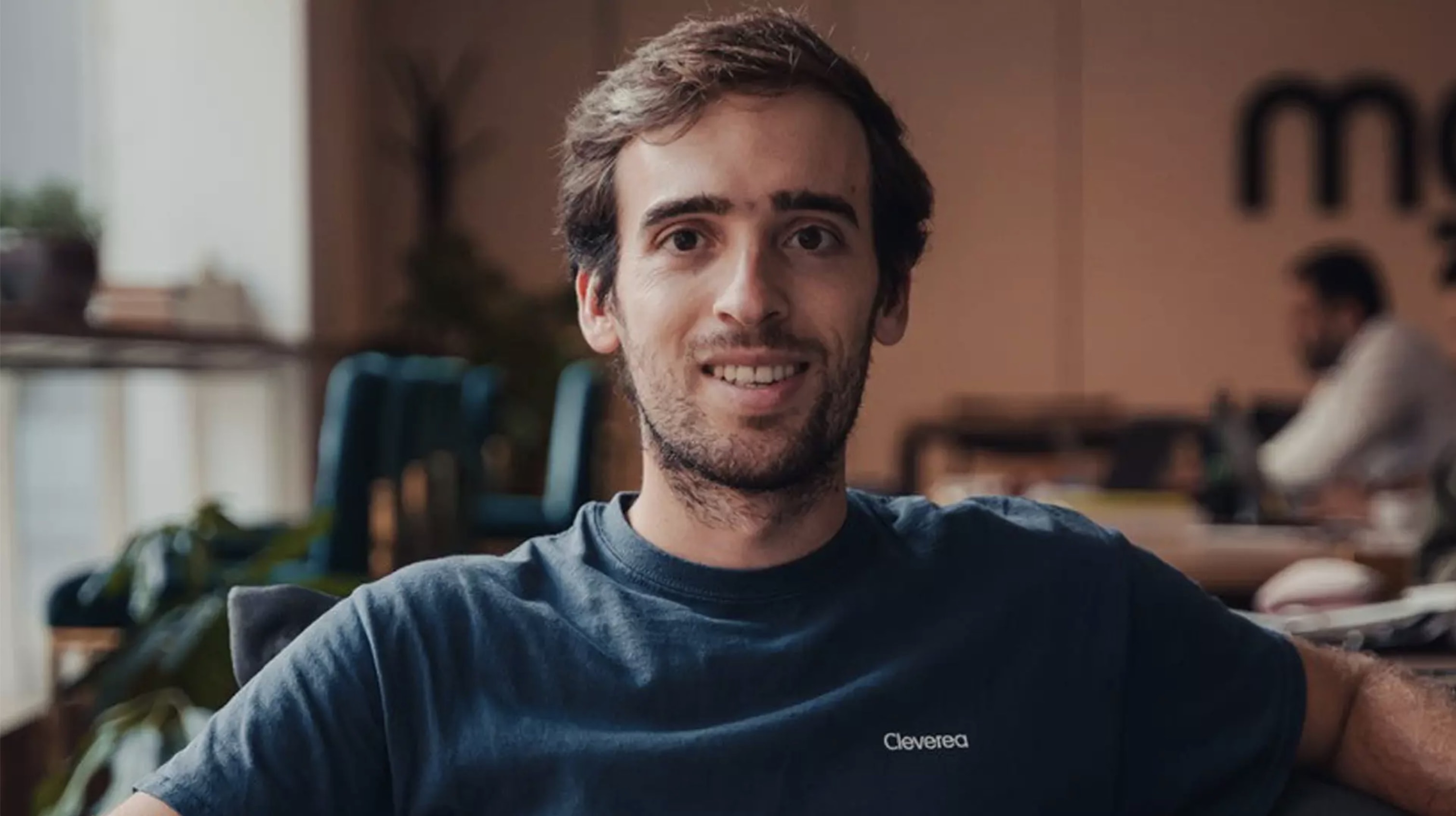 Javier Bosch, co-fundador de Cleverea, sonriente con camiseta azul, sentado en un entorno de oficina moderna con plantas y mesas de trabajo al fondo.