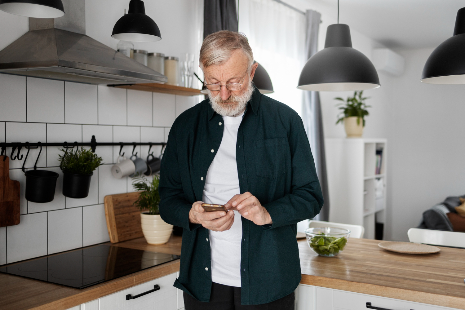 Hombre mayor con barba y gafas, usando un teléfono móvil en una cocina moderna con plantas y utensilios colgados.