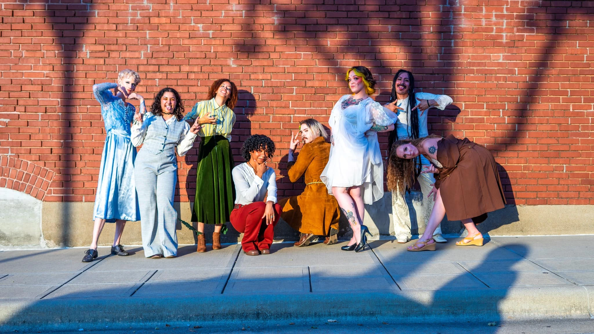 8 models standing along a brick wall in 70's Victorian-Bohemian style outfits.