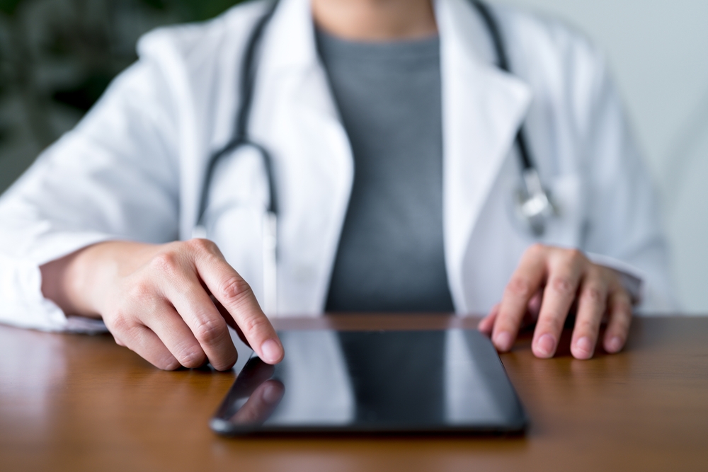 Healthcare professional in white coat using a digital tablet on a wooden table.