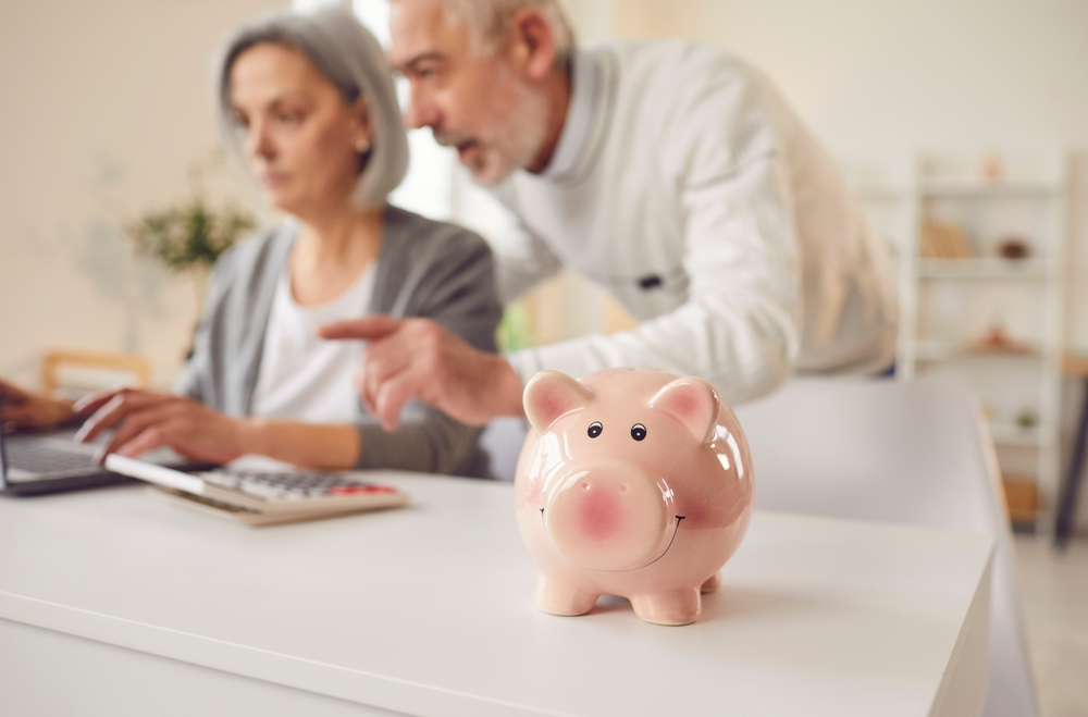 Smiling pink piggy bank on a white table with an elderly couple using a laptop and calculator in the background.