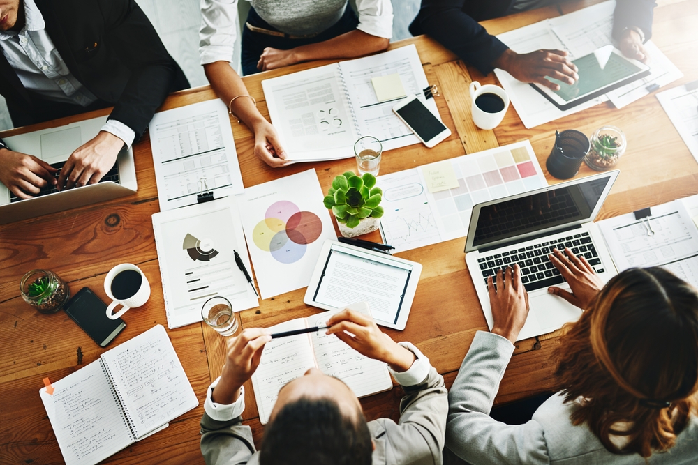 Overhead view of a diverse group of people collaborating around a wooden table with laptops, tablets, documents, charts, notebooks, coffee cups, and potted plants.