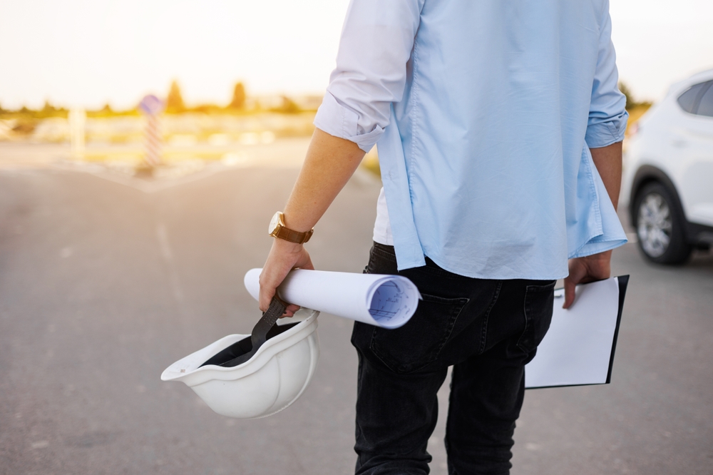 Person in blue shirt holding a white hard hat and rolled-up construction blueprints on a street near a white car.