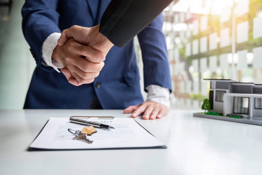 Two people shaking hands over a signed contract with house keys and a model house on the table.