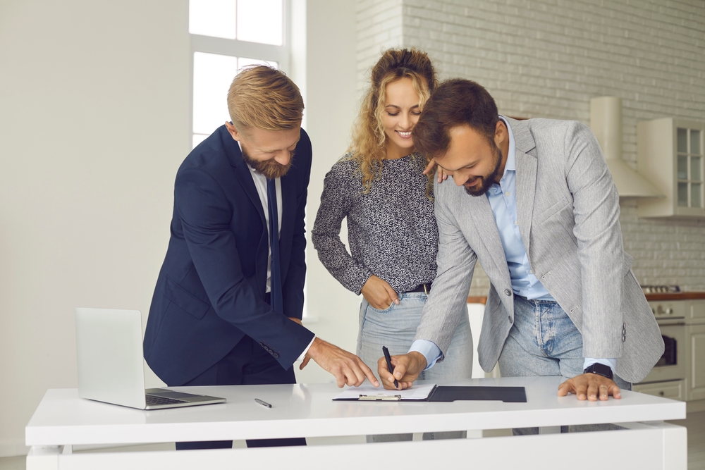 Two men and a woman in business attire reviewing and signing a document on a white table in a bright office.
