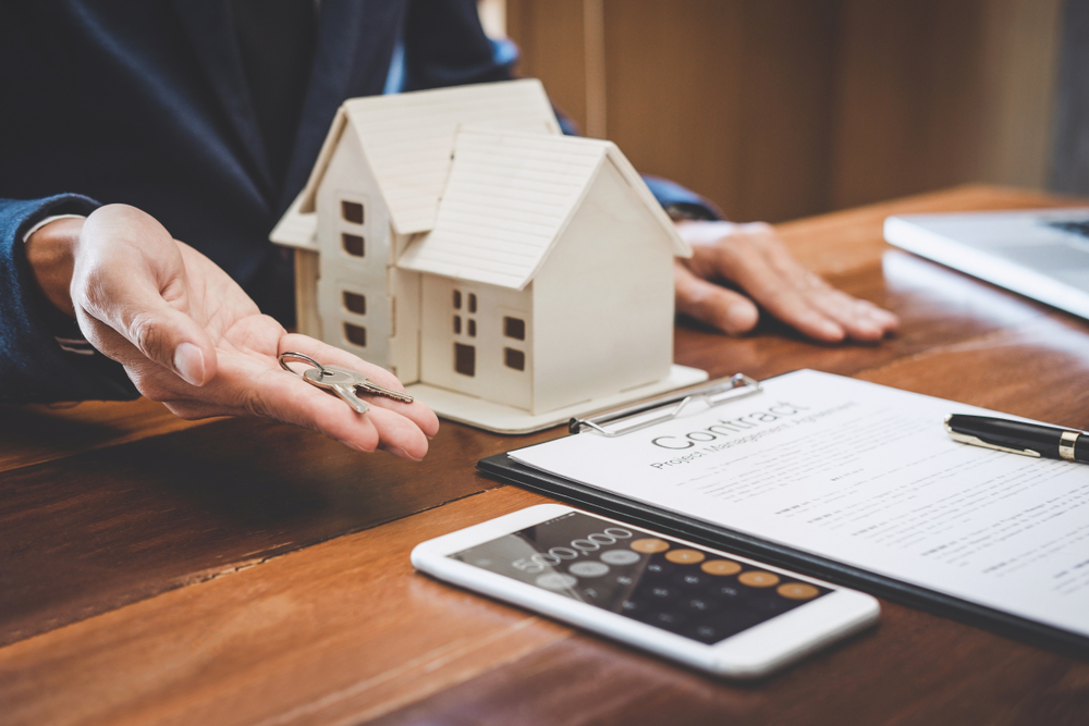 Person holding house keys near a wooden model house with a contract, pen, and calculator on the table.