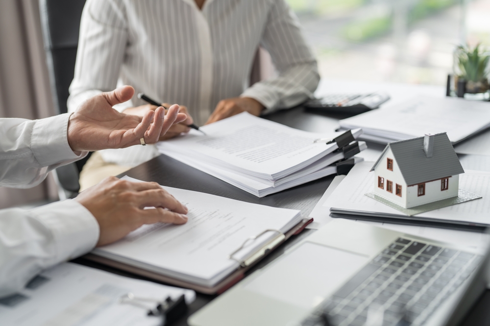 Two people discussing documents at a desk with a small house model and a laptop, indicating real estate or contract review.