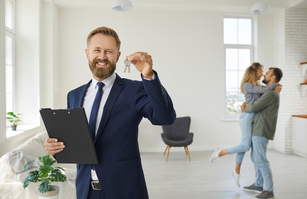 Smiling real estate agent holding a clipboard and keys with a happy couple embracing in the background of a bright, empty apartment.