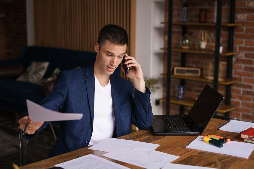Young man in a blue blazer talking on a phone while holding documents at a wooden desk with a laptop and scattered papers.