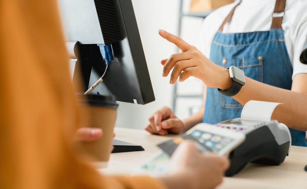 Barista in denim apron using a computer while a customer holds a credit card near a payment terminal.