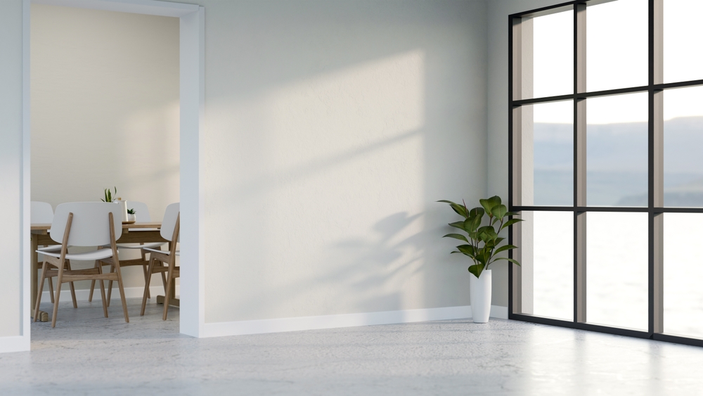 Minimalist room with large black-framed window, white walls, concrete floor, a green plant in white pot, and a dining area with wooden chairs and table in the adjacent room.