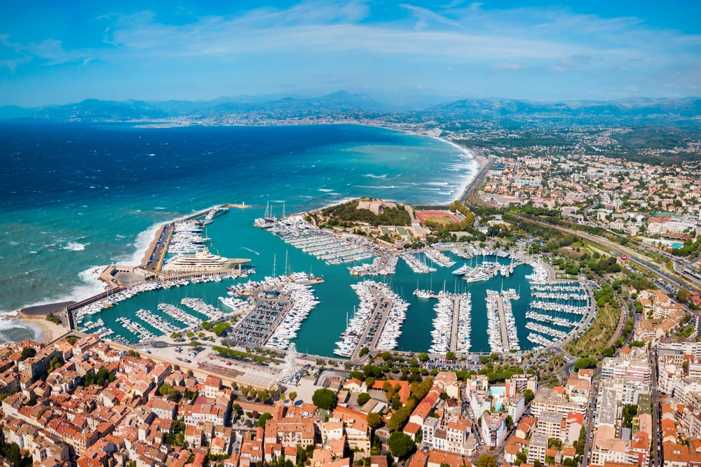 Aerial view of a coastal marina with numerous yachts docked, a surrounding town with red-roofed buildings, and a curved coastline with blue ocean stretching into the distance.