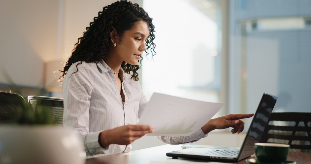 Woman with curly hair reviewing a paper and pointing at a laptop screen in a bright office.