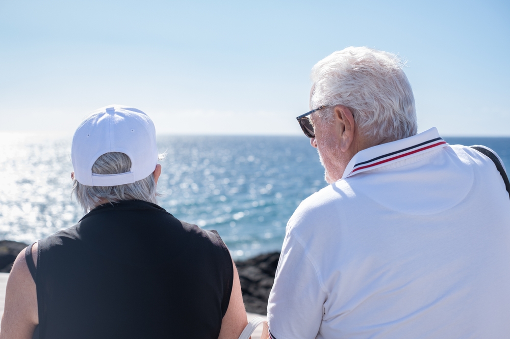 Elderly couple sitting by the ocean under clear blue sky, viewed from behind.