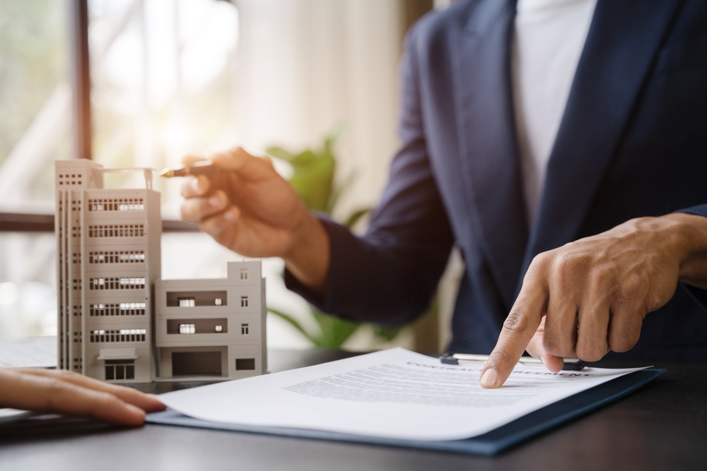 Person in suit pointing at a document on a clipboard with a miniature building model on the table.