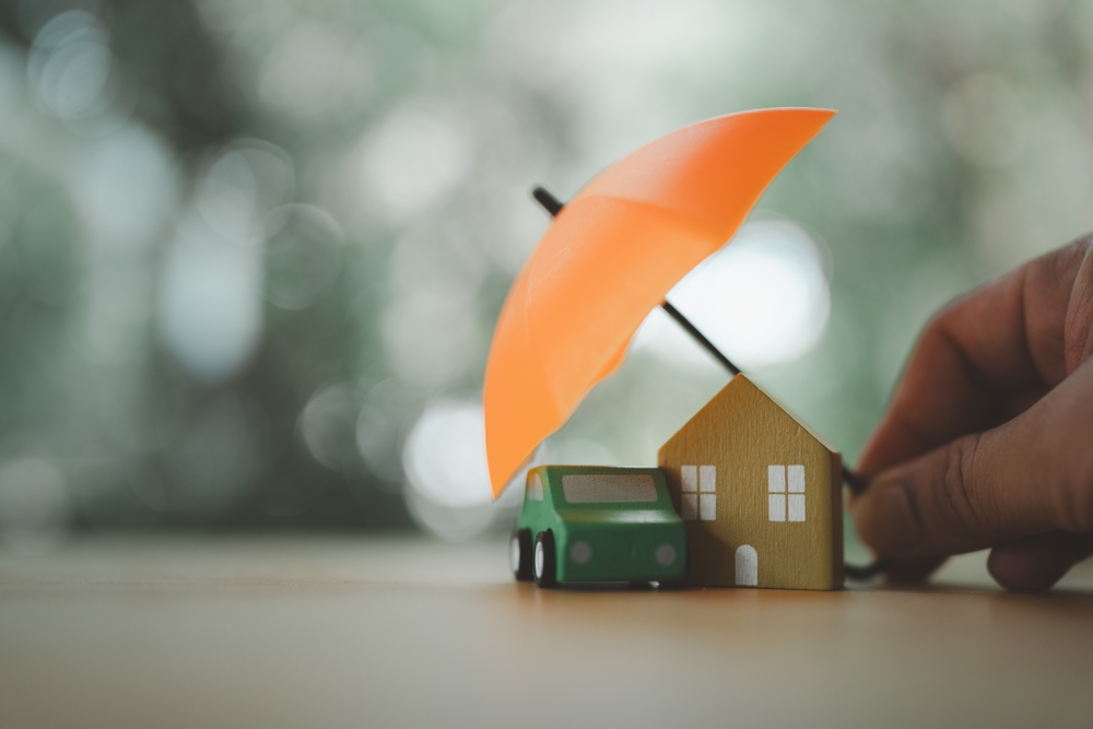 Hand holding an orange umbrella protecting a small wooden house and green toy car on a blurred background.