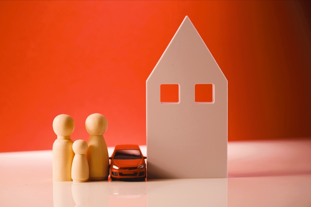 Wooden figurines representing a family with a toy car next to a white house model against an orange background.