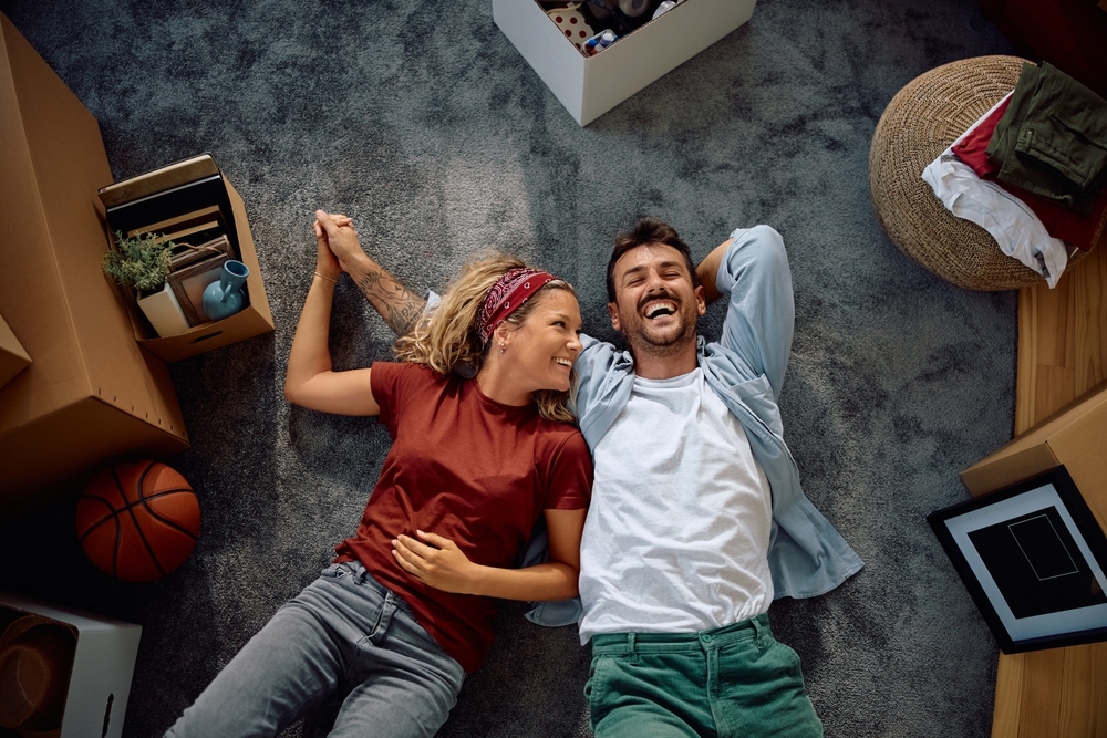 Smiling couple lying on a carpeted floor holding hands, surrounded by moving boxes and household items.