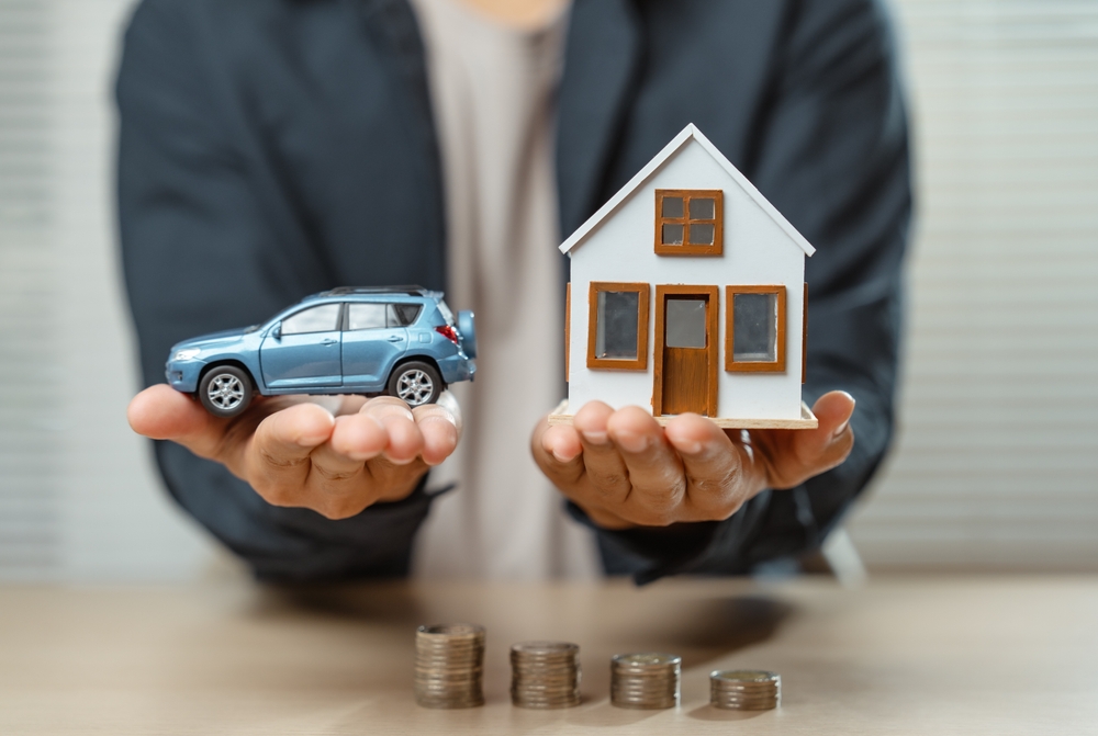 Person holding a miniature blue car in one hand and a model house in the other, with stacked coins on the table below.