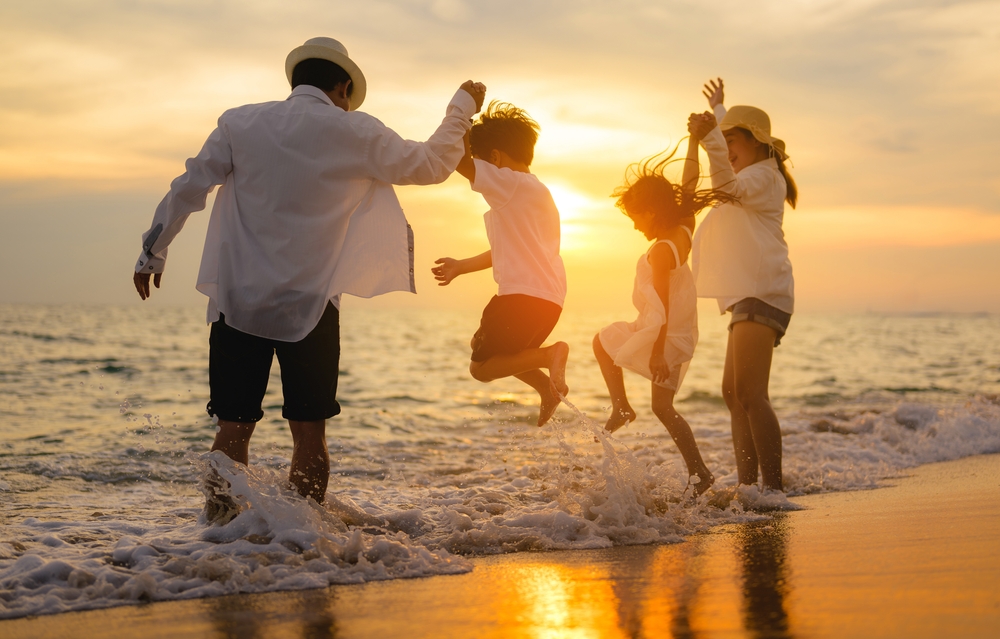 Family of four holding hands and jumping over waves on a beach at sunset.