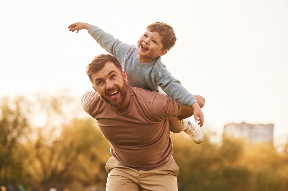 Smiling man giving a piggyback ride to a happy young boy with arms outstretched in a park at sunset.