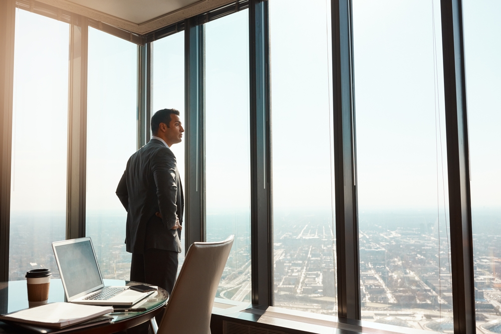 Businessman in suit standing with hands on hips, looking out large office windows over a cityscape.