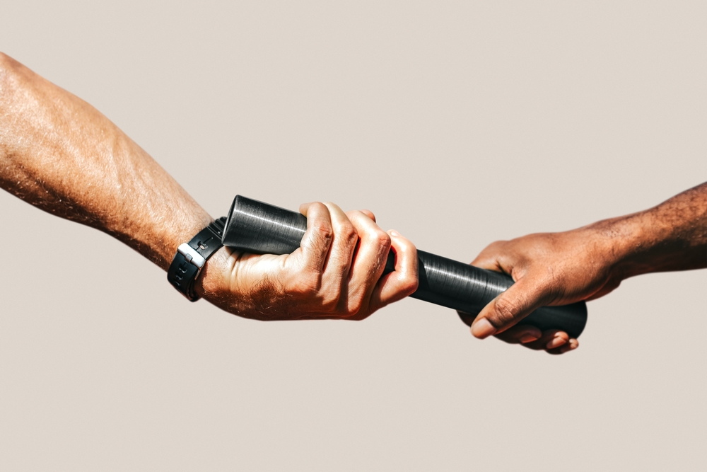 Two hands exchanging a black relay baton against a neutral background.