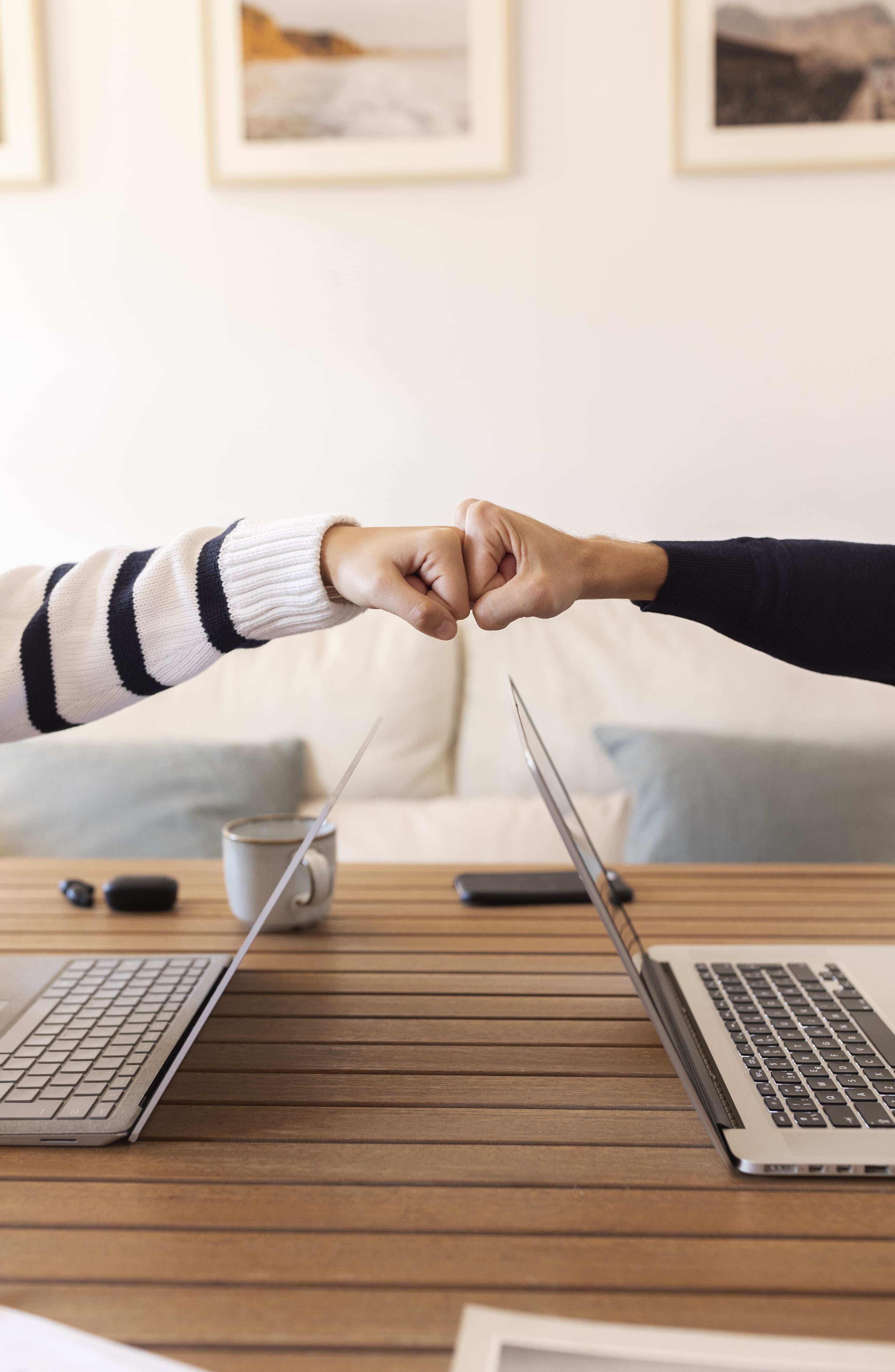 Two people fist bumping over a wooden table with two open laptops, a coffee cup, and a smartphone.