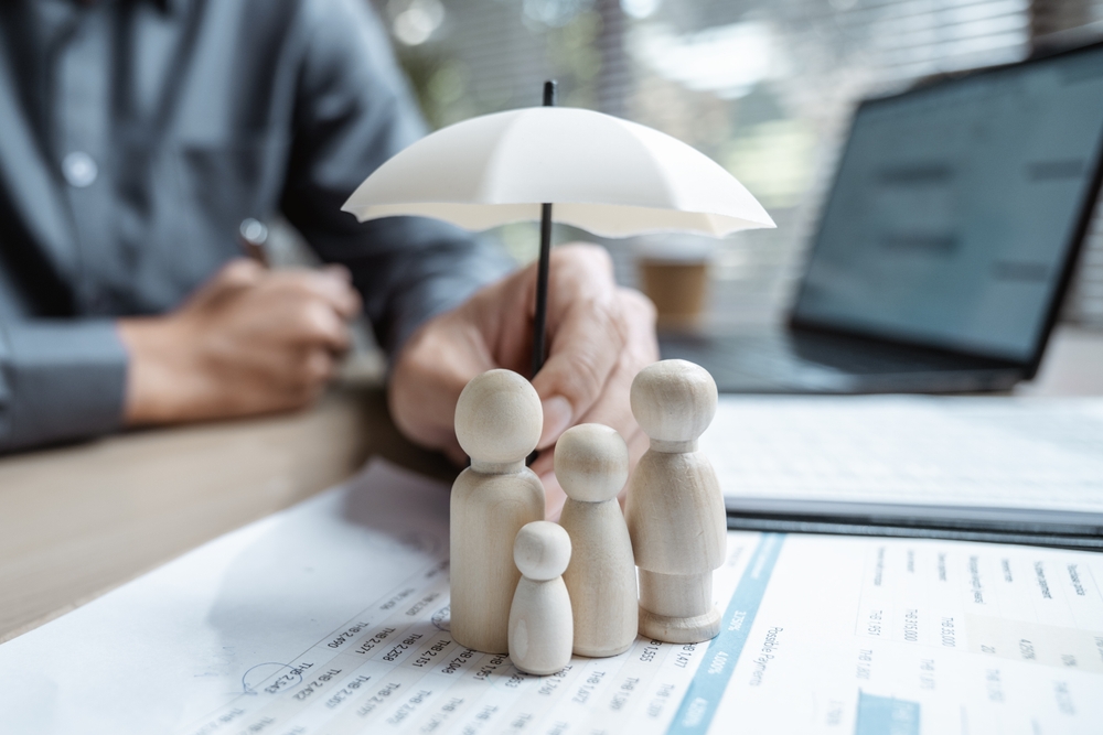 Wooden family figurines protected by a small white umbrella held by a person over financial documents on a desk.
