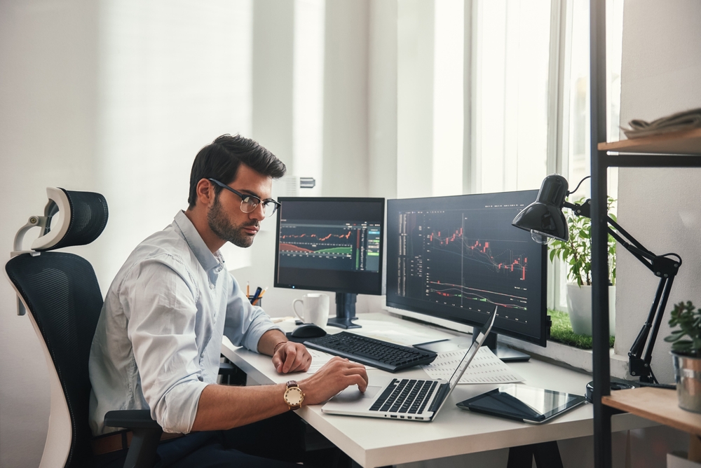 Man in glasses working on a laptop at a desk with two monitors displaying stock market charts.